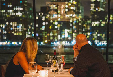 An intimate dining scene with two people at a table by a window overlooking a city at night. Their backs to the camera, they appear in silhouette against the sparkling urban backdrop. A bottle and glasses of wine on the table complete the cozy setting.