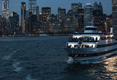 A ferry boat moves across the foreground with the evening lights of a city skyline in the background. The water is slightly choppy, and the sky above is dusky, suggesting either dawn or dusk. The city's buildings are lit from within, twinkling in the dim light.