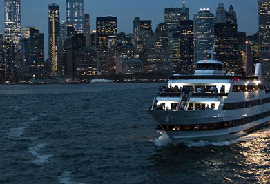 A ferry moves across the water in the early evening with a backdrop of a city skyline with illuminated buildings under a dusky sky.