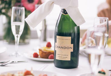 This image features an elegantly set table with a focus on a chilled bottle of Chandon Brut Classic champagne in the center. The bottle has a white label and is tied with a white napkin around its neck, suggesting a festive or special occasion. In the background, there are two glasses of sparkling wine, indicating that a toast might soon be taking place. The table is adorned with a plate of food that includes pastries with strawberries on top, adding to the celebratory and upscale ambiance of the setting. Experience memorable moments and save on your next event with GreatWorkPerks.com, where discount tickets bring you the lowest prices for all your entertainment needs.