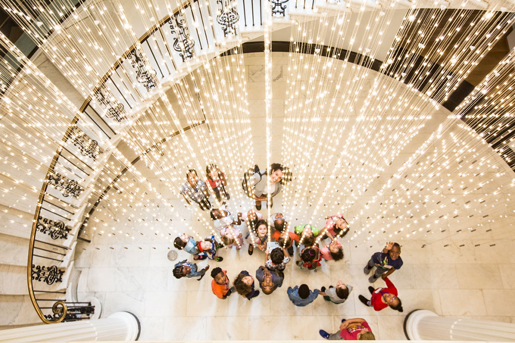 An overhead view of a bustling lobby adorned with a dazzling array of hanging lights creating a canopy of warm illumination. Below, visitors stand and walk, some gazing upward, the grandeur of the staircase railing adding to the scene's elegance.