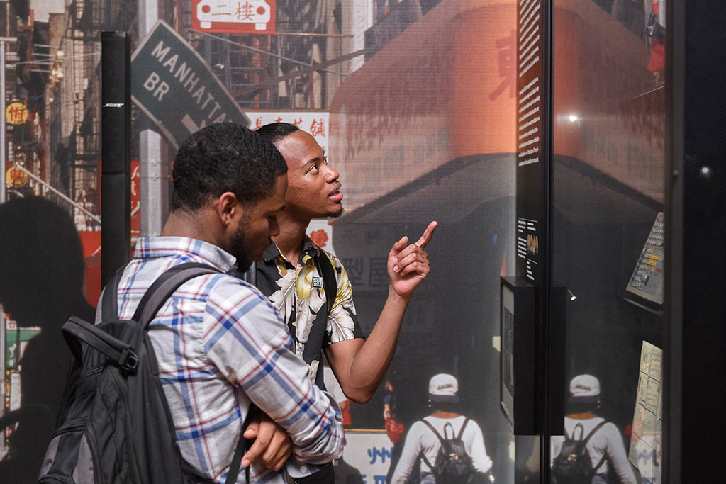 Two individuals are engaged in viewing an exhibit with urban-themed imagery, including a Manhattan Bridge sign. One person gestures with raised finger, likely explaining a point of interest.