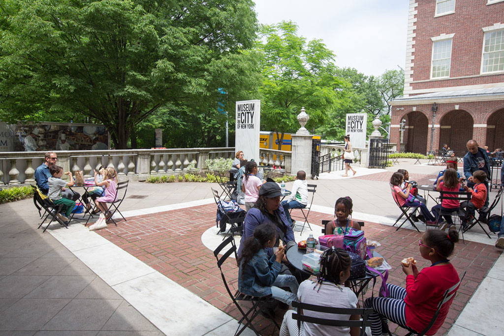 Outdoor setting with multiple people sitting at round tables enjoying a casual gathering. Trees shade parts of the area, and a brick building with archways is visible in the background. There is a sign with  MUSEUM CITY OF NEW YORK  in the view. The gathering has a relaxed atmosphere in a city space.