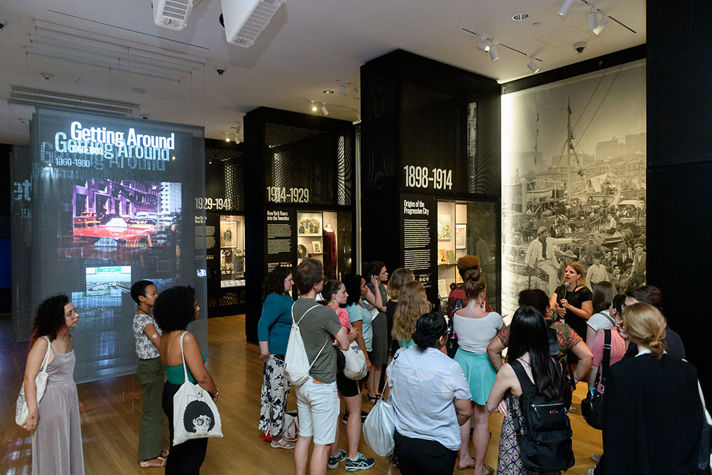 A diverse group of visitors participating in a guided tour inside a museum exhibit about urban transportation history, with displays spanning from 1898 to 1980 and including large photographs and informative texts.