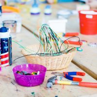 This image features a vibrant and dynamic arts and crafts workspace. At the center is a wicker basket overflowing with a tangle of colorful yarn or string. To the left lies a container of various paints, including a large white bottle and a smaller blue tube, possibly acrylics. Nearby, a pink plastic bowl is filled with a collection of crayons, hinting at a playful and creative setting. In the foreground, an assortment of tools can be observed, such as pliers and a pair of orange-handled scissors, suggesting a variety of crafting activities take place here. The work surface beneath these items appears well-used, adorned with splatters of paint and marks from previous projects. Just like the multitude of supplies in this arts and crafts image, FunEx.com offers an array of options to cater to your entertainment needs, all at the lowest prices—ensuring you can enjoy more fun for less.