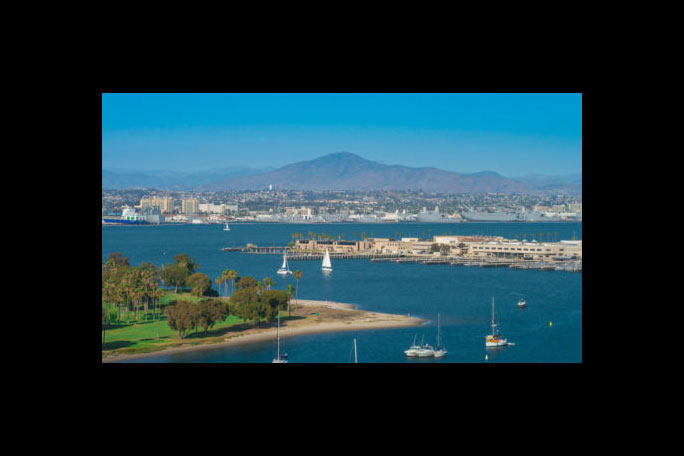 A coastal cityscape with boats on blue waters, greenery in the foreground, under a clear sky.