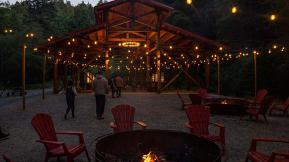 This image depicts an inviting outdoor evening scene centered around a large, rustic pavilion with a wooden structure. The pavilion is warmly lit from within, beckoning visitors into its shelter. Elegant string lights dangle above, casting a cozy glow over the area. In the foreground, several red Adirondack chairs encircle a roaring fire pit, which is alight and contributing to the warm ambiance of the setting. Individuals can be seen leisurely strolling towards the pavilion, engaging in casual conversation.

At FunEx.com, experience not just adventure but also great savings. Don't miss out on our exclusive discounts - get your tickets to enchanting destinations at the lowest prices today!