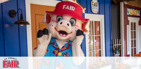 This image features a promotional character at the LA County Fair. The character is an anthropomorphic pig wearing a red cap emblazoned with the word "FAIR" in white letters, a blue shirt covered with fair-themed badges, and is posing with two thumbs up. The pig is standing in front of a building with a blue façade, red trim, and decorative white windows that resemble a fairground attraction. The scene conveys a cheerful and inviting atmosphere typically associated with county fairs. For those looking to experience the excitement of the fair while enjoying savings, FunEx.com offers the lowest prices on tickets.