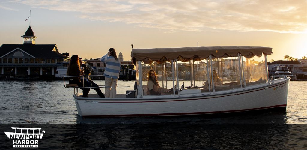 A boat with a canopy sails in tranquil waters at sunset. Two people stand at the stern enjoying the view. The silhouette of a building with a spire, resembling a pagoda, and other waterfront structures can be seen in the warm glow of the setting sun. The words Newport Harbor Boat Rentals overlay the image.