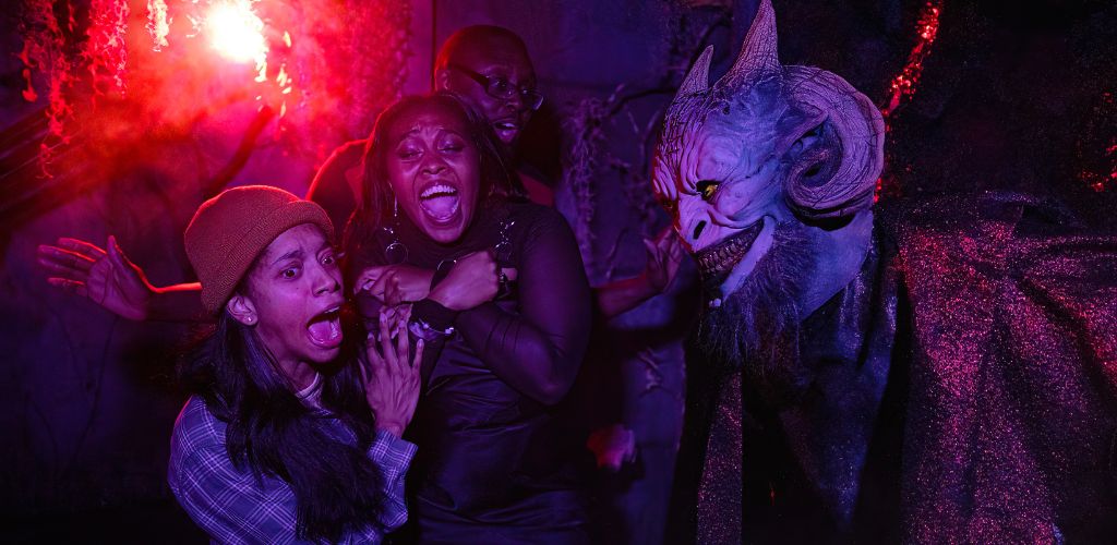 Image shows a thrilling scare experience with three individuals expressing fear and surprise. A person in a monster costume startles them amidst a backdrop illuminated by a red flare. Emotions run high in this vividly captured moment.