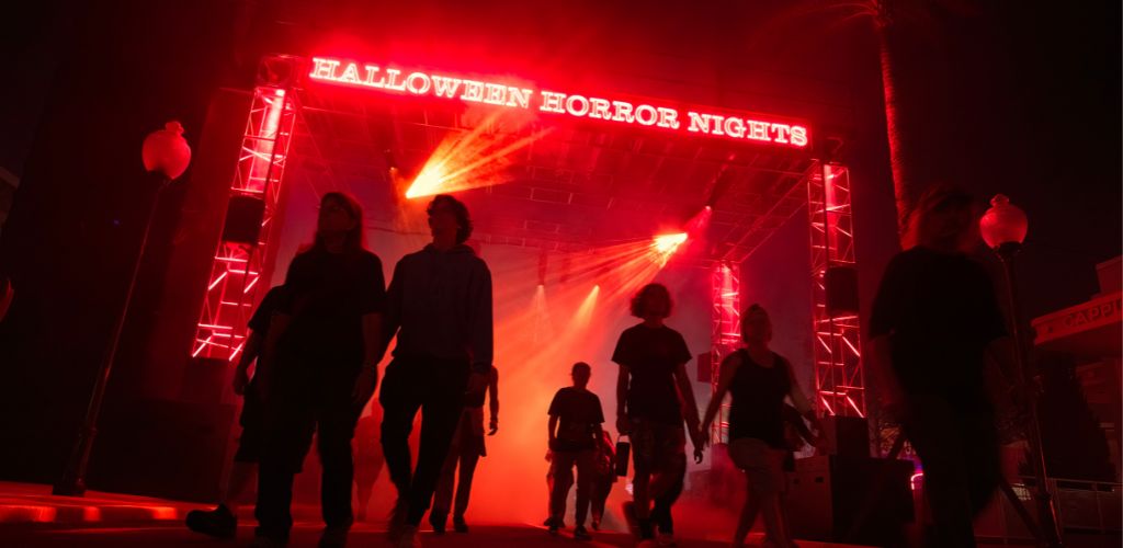 Image shows a crowd of people walking towards an entrance with a sign reading 'Halloween Horror Nights'. The atmosphere is eerie with a red and dark ambiance created by theatrical fog and lighting. The excitement of an evening filled with spooky entertainment is palpable.