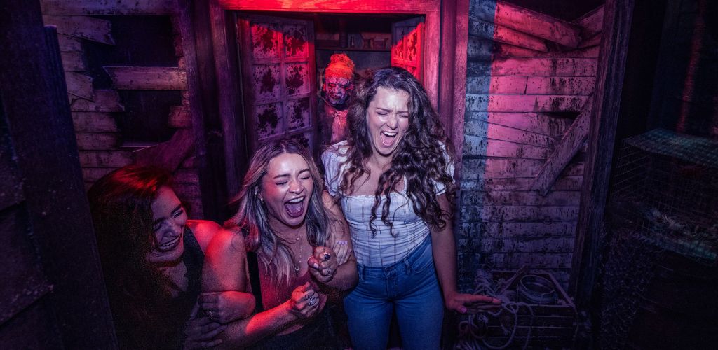 Three people express fear and amusement as they exit a dimly lit space resembling a haunted house. A person with special makeup, suggesting a zombie-like character, lurks in the background. Moody lighting with blues and reds enhances the eerie atmosphere.