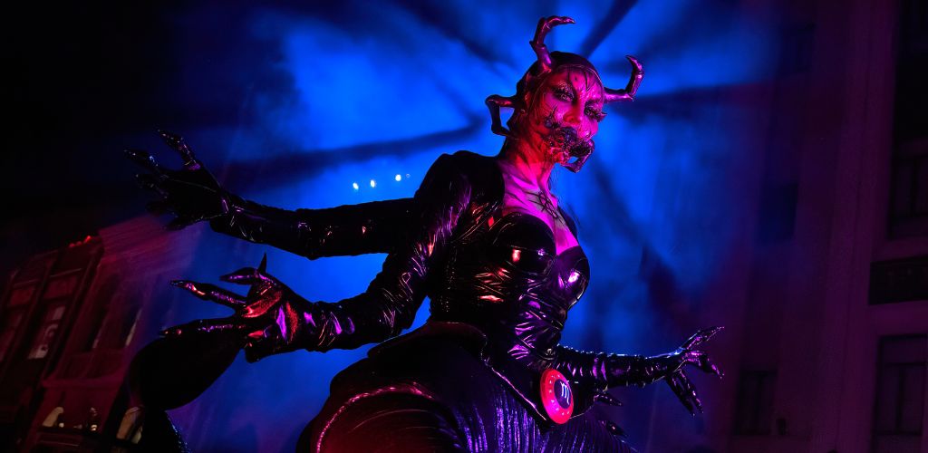 Image depicts a person in an elaborate devil costume with horns, under blue theatrical lighting. The costume features shiny black material, and the individual appears poised dramatically against a blurred background suggestive of a night-time setting.