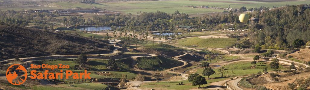 Panoramic view of a spacious outdoor animal park with winding paths amidst rolling hills and varied greenery. A tethered, yellow balloon is visible in the distance, against a clear blue sky. The location's logo featuring an animal silhouette is on the bottom left.