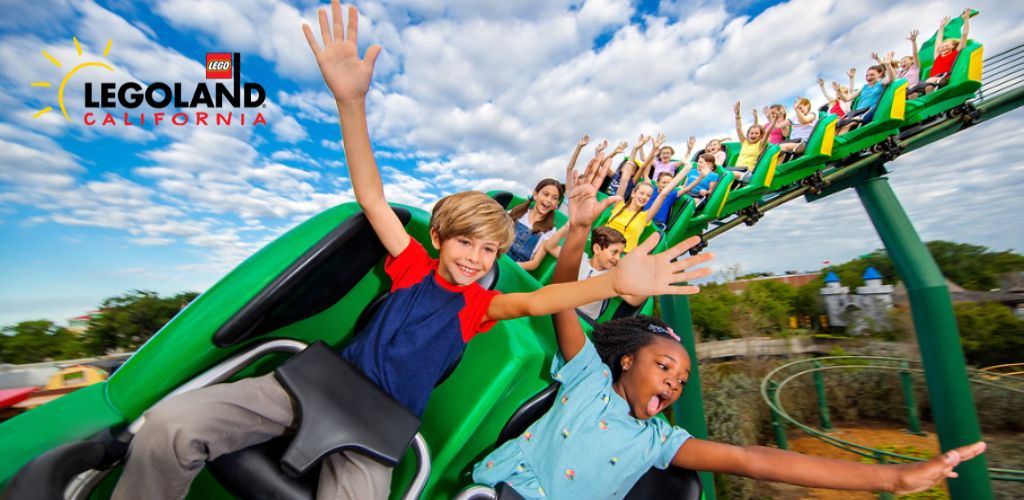 Image Description: Excited children and adults are seen riding a green roller coaster with their arms raised in joy. They are seated in pairs along the coaster train, which is adorned with the 'LEGOLAND California' logo. The backdrop features a bright blue sky with fluffy clouds and bits of lush greenery at the ride's base. The sense of motion and fun is palpable in this dynamic amusement park setting.