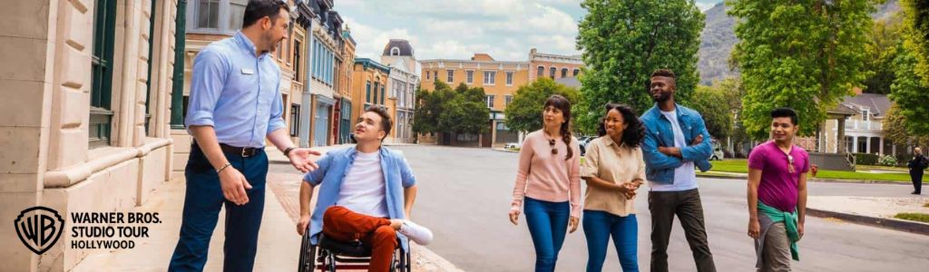 Image shows a diverse group of visitors enjoying a tour at the Warner Bros. Studio. A guide in a blue shirt and beige pants is talking to a young visitor in a wheelchair. Other guests walk casually alongside, engaging with the surroundings on a sunny day, with studio buildings and mountains in the background.