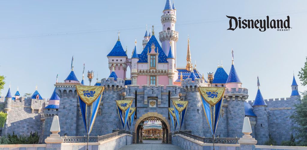 Image showing the iconic Sleeping Beauty Castle at Disneyland Resort on a sunny day. The sky is clear and blue, complementing the castle's pastel pink and blue turrets adorned with golden accents. Blue and gold banners hang along the stone castle walls leading to an arched entrance. The Disneyland Resort logo is in the upper right corner.
