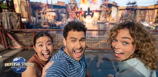 Three excited friends take a selfie with a large explosion in the background at Universal Studios Hollywood. They're sporting wide smiles, capturing the thrill of the attraction. The Universal Studios logo is visible in the corner.