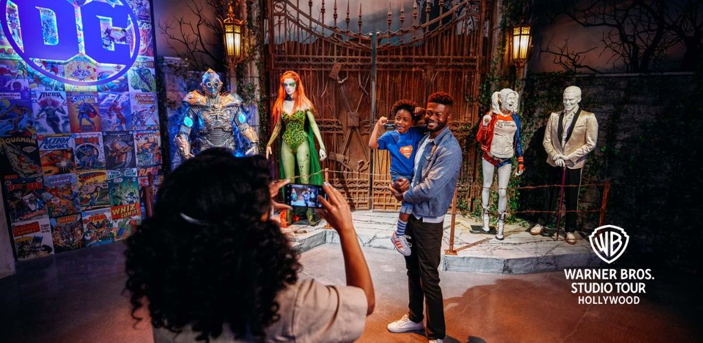 Visitors pose for a photo at Warner Bros. Studio Tour Hollywood in a DC-themed exhibit. Lifesize figures of iconic characters surround them while a person in the foreground takes their picture. The setting includes a large DC logo and a backdrop featuring comic book covers.