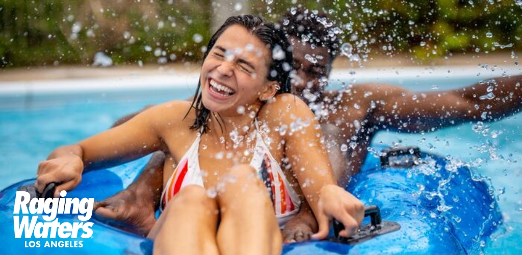 Image features two individuals enjoying a water ride. A person in the foreground is laughing, surrounded by water splashes, while riding an inflatable blue tube. In the background, there's another rider, partially obscured. Text on image reads 'Raging Waters Los Angeles'. The atmosphere is vibrant and joyful.