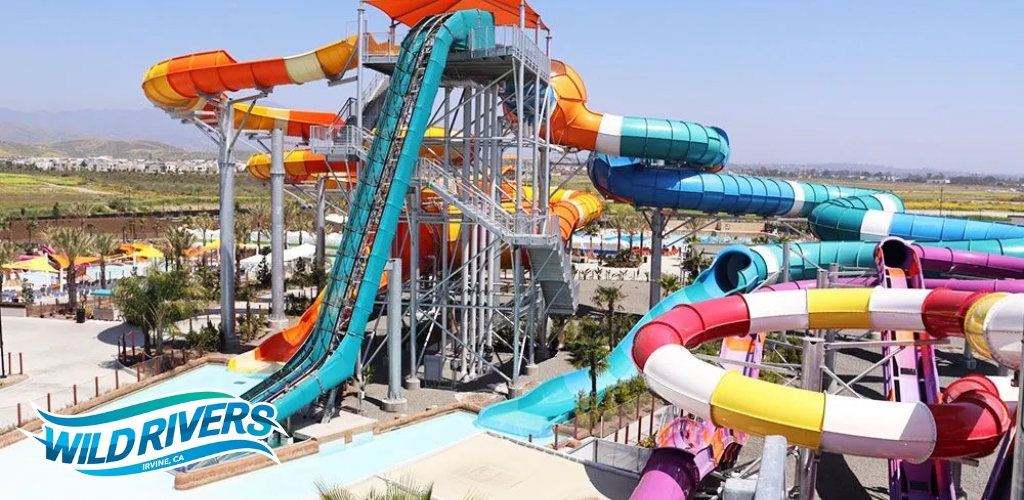 A vibrant water park scene featuring a series of intertwined, colorful water slides in blue, orange, green, and pink. A metal staircase structure provides access to the slide entrances. Below, a pool awaits sliders. Palm trees and lounge areas are visible in the background under a clear sky. The logo on the image indicates 'Wild Rivers' in Irvine, CA.