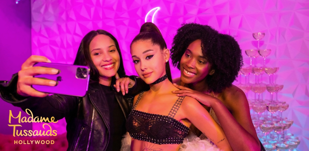 Image displays two individuals posing for a selfie with a wax figure at Madame Tussauds Hollywood. The person on the left holds the smartphone, embracing the wax figure in the middle, while the person on the right smiles and rests an arm on the figure's shoulder. They stand before a geometric-patterned purple backdrop, with a tower of champagne flutes nearby. The setting has a vibrant, celebratory ambiance.