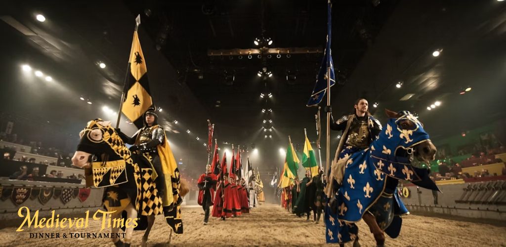 Medieval Times Dinner and Tournament scene with knights and horses parading in an arena. The foreground shows a knight in blue armor on a matching decorated horse, holding a blue flag with fleur-de-lis. Spectators are seated in the background.