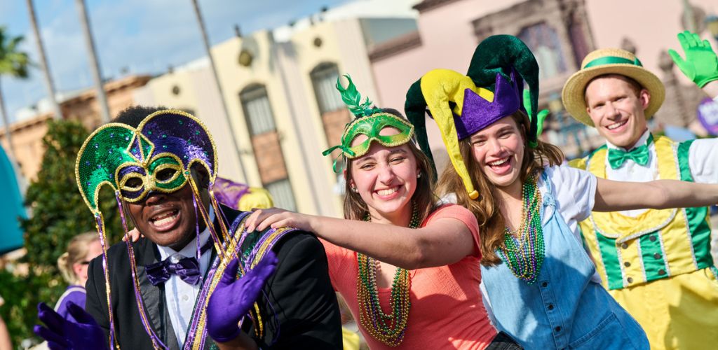 A lively group of people celebrate in vibrant Mardi Gras costumes. They wear colorful masks, hats, and beads, smiling and dancing in a parade setting. The background shows palm trees and festive buildings, reflecting a joyful and energetic atmosphere perfect for an exciting carnival or festival event.