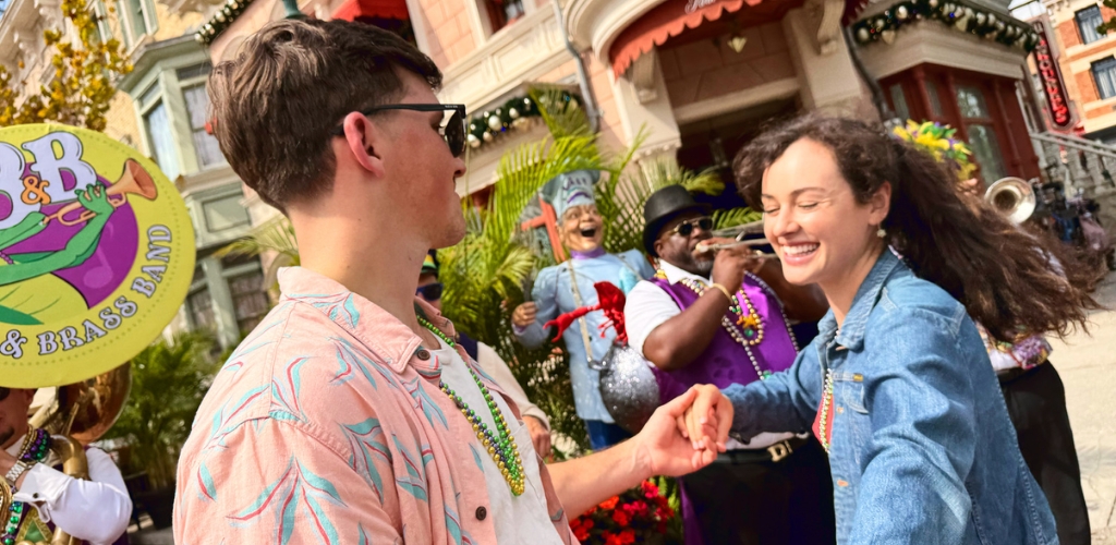 A joyful couple dances in the street during a lively festival. The man wears sunglasses and a colorful shirt, while the woman, smiling widely, wears a denim jacket. They hold hands surrounded by a vibrant brass band with musicians in festive attire. The background features charming buildings adorned with colorful decorations, capturing the essence of a fun, lively outdoor celebration.