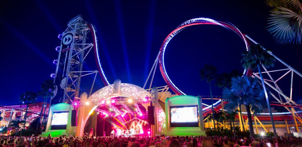 A vibrant amusement park scene at night with a live concert on an illuminated stage in the foreground. The backdrop features a towering roller coaster with loops and bright lights against a dark sky. Palm trees and a crowd of spectators complete the lively atmosphere, creating an exciting and energetic environment ideal for entertainment and fun.