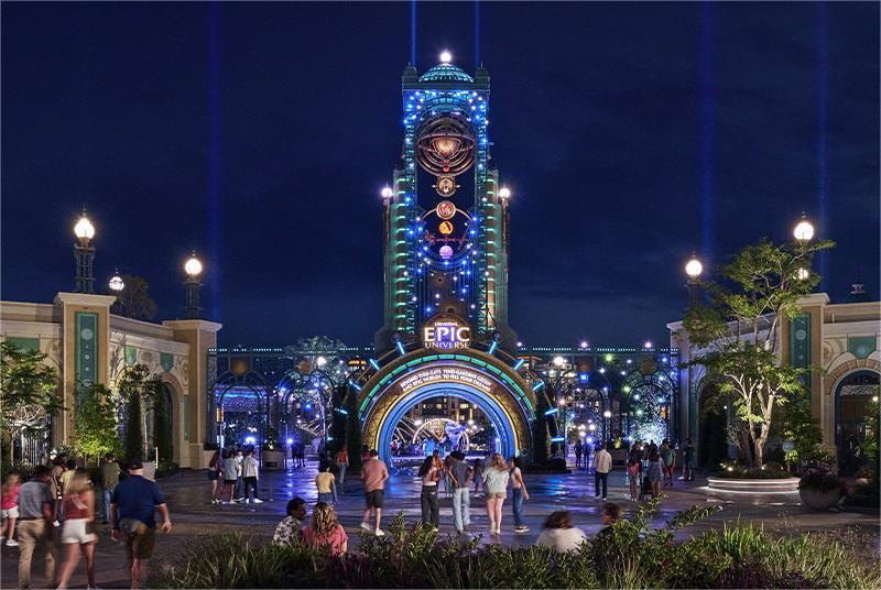 A towering steampunk portal aglow in sapphire and gold lights under a deep night sky, with guests strolling across the glistening plaza toward the radiant “Epic Universe” arch.