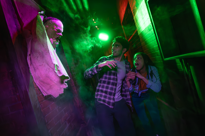 Two people stand in a dark hallway illuminated by eerie green lights, creating a spooky Halloween atmosphere.