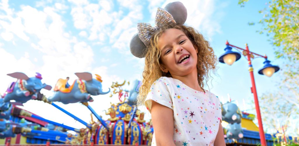 A smiling girl with curly hair and mouse ears headband enjoys a carousel ride at an amusement park on a sunny day