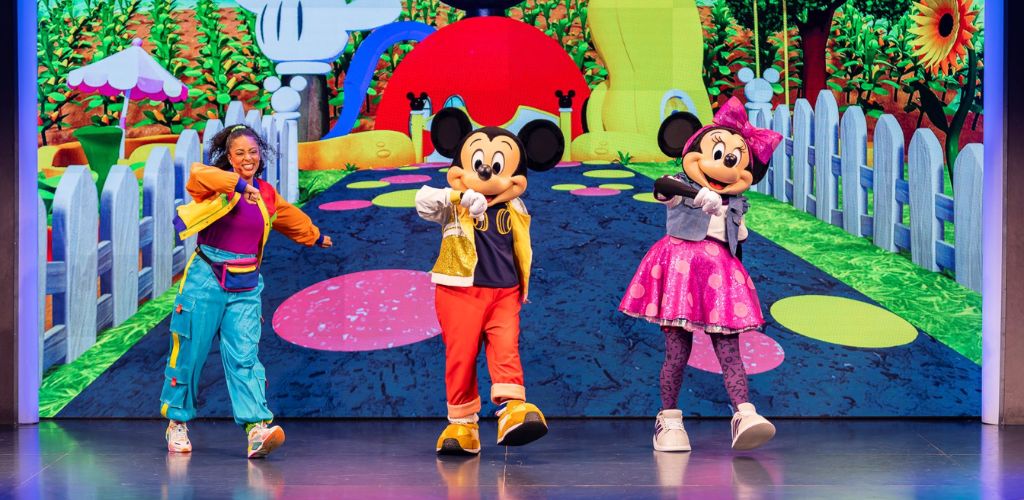 Three entertainers in colorful costumes dance in front of a vibrant animated background featuring a whimsical garden scene with plants and characters.
