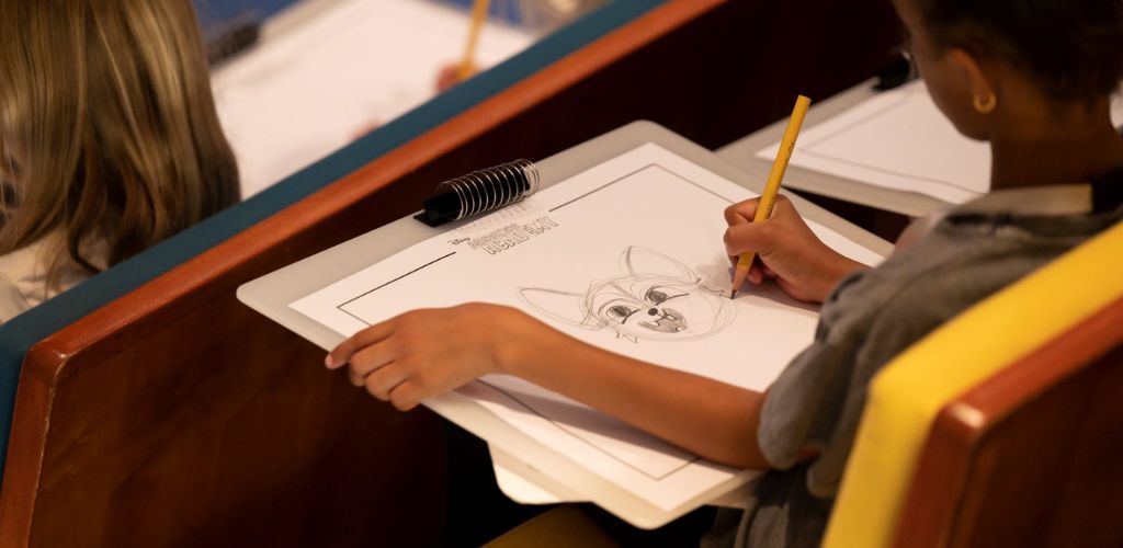 A child sitting at a desk drawing a cartoon character on paper with a yellow pencil.