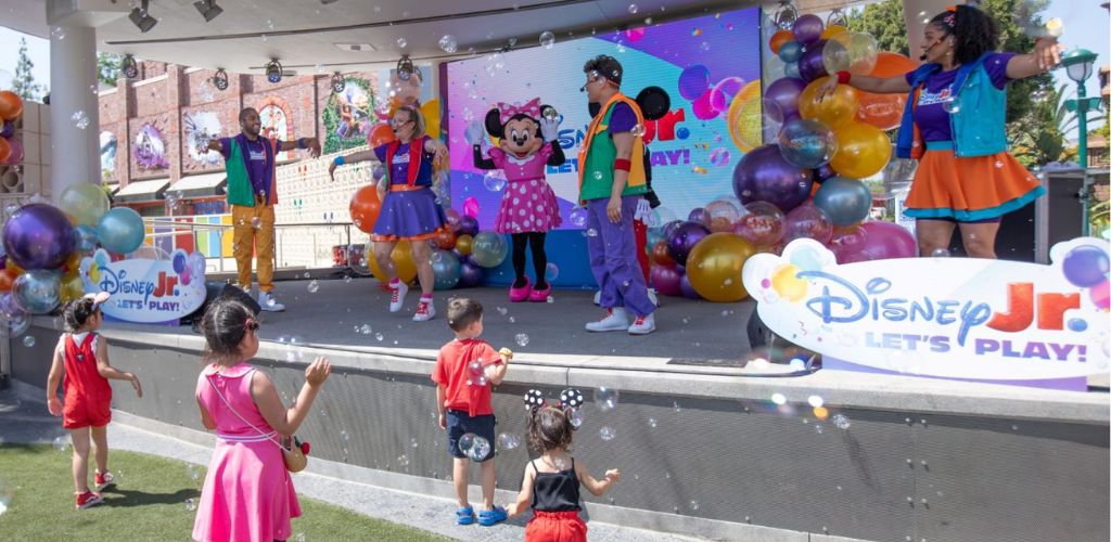 Children watching a colorful stage show featuring Disney characters and balloons at an outdoor event for kids.