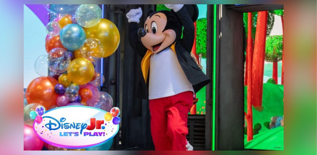 Mickey Mouse character posing at Disney Jr Let's Play event with colorful balloons and vibrant background.