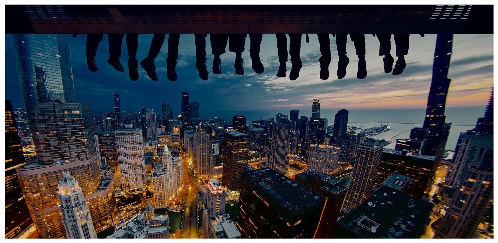 People sitting on a ledge overlooking a city skyline at dusk with buildings illuminated and the sky transitioning from sunset to night.