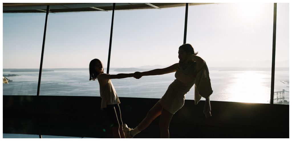 A woman and girl holding hands and smiling indoors with large windows overlooking a body of water and distant islands during sunset.
