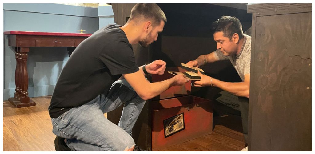 Two men are exchanging items inside a wooden cabinet under a desk in a room with wooden flooring.