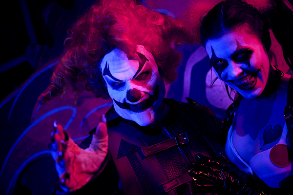 Two people dressed as scary clowns with painted faces and dark costumes, smiling and posing in a dimly lit, colorful nightclub scene
