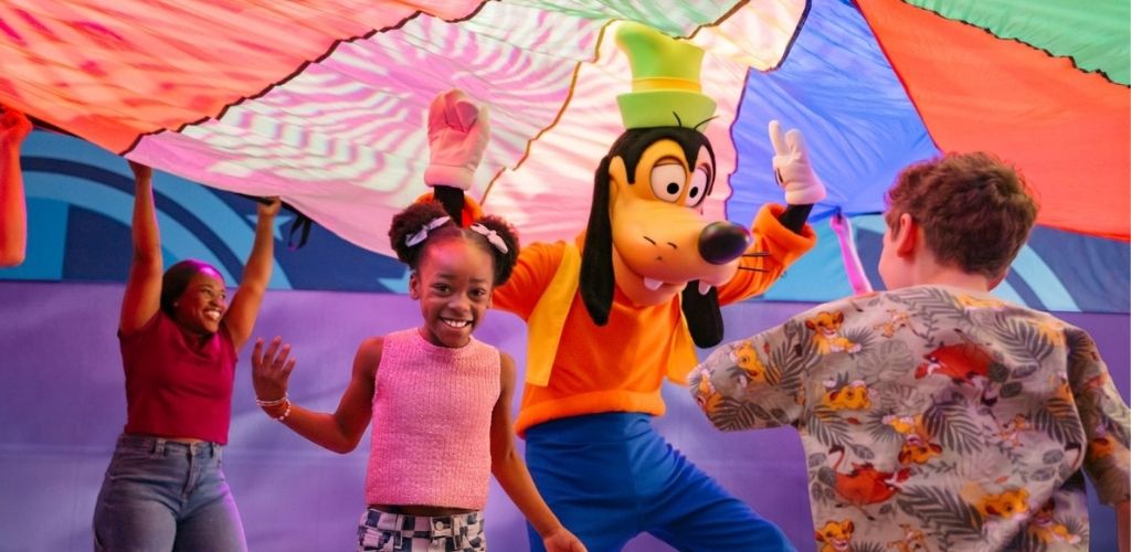 Children smiling and playing with a character in a large colorful parachute outdoors