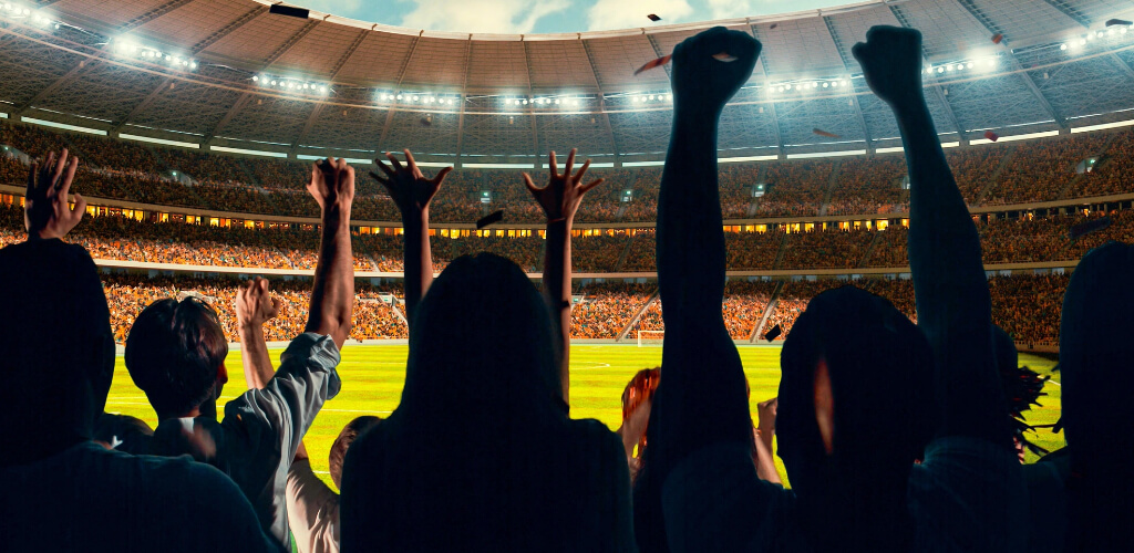 Silhouettes of cheering fans with raised arms inside a packed stadium during a sporting event.