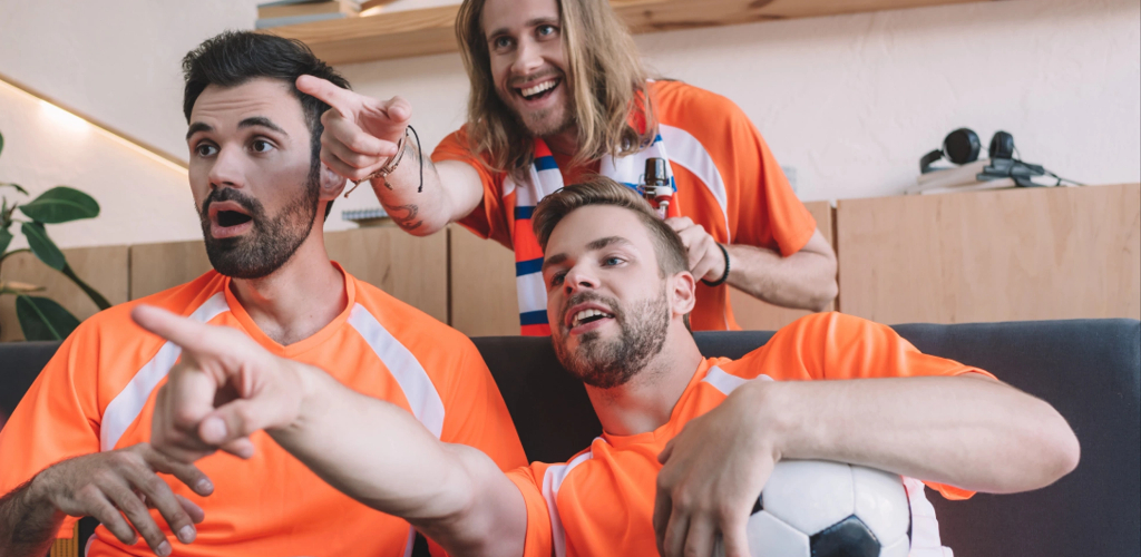 A group of excited fans in orange jerseys cheering and reacting while watching a soccer match together.
