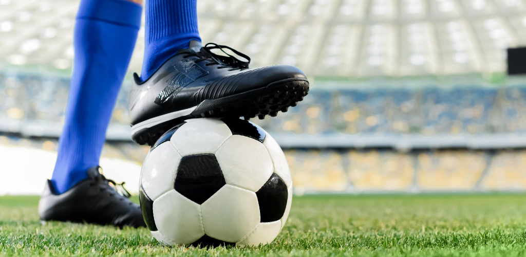 A soccer player in blue socks and black cleats resting one foot on a black-and-white soccer ball on a stadium field.