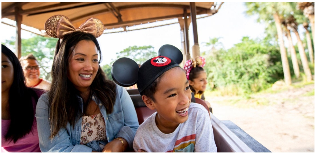 Group of smiling children and adults enjoying a fun ride at an amusement park with Disney themed hats and accessories in a sunny outdoor setting