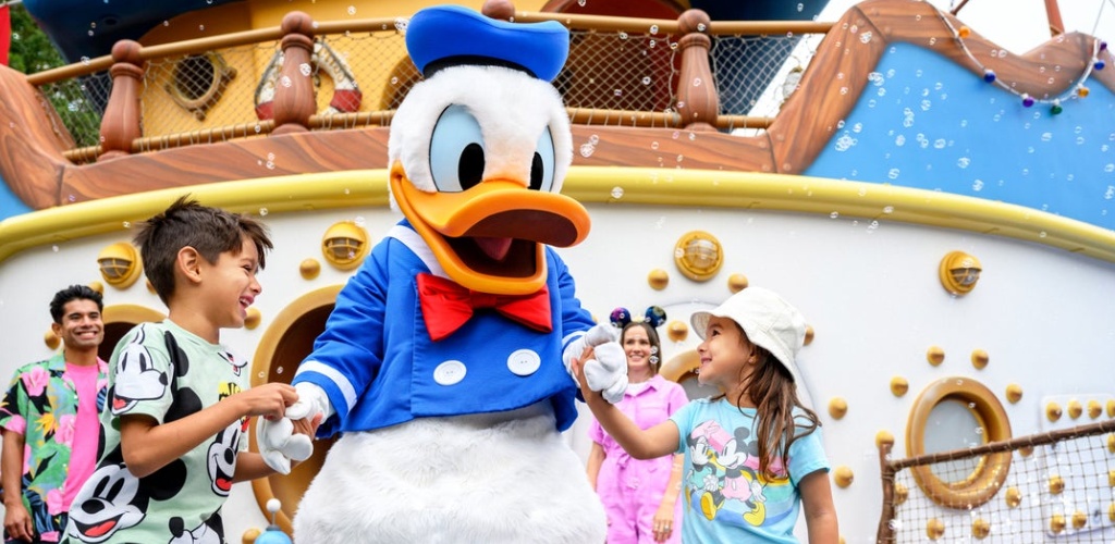 Children smiling and holding hands with a large Donald Duck mascot at an outdoor amusement park with a decorated themed ship in the background