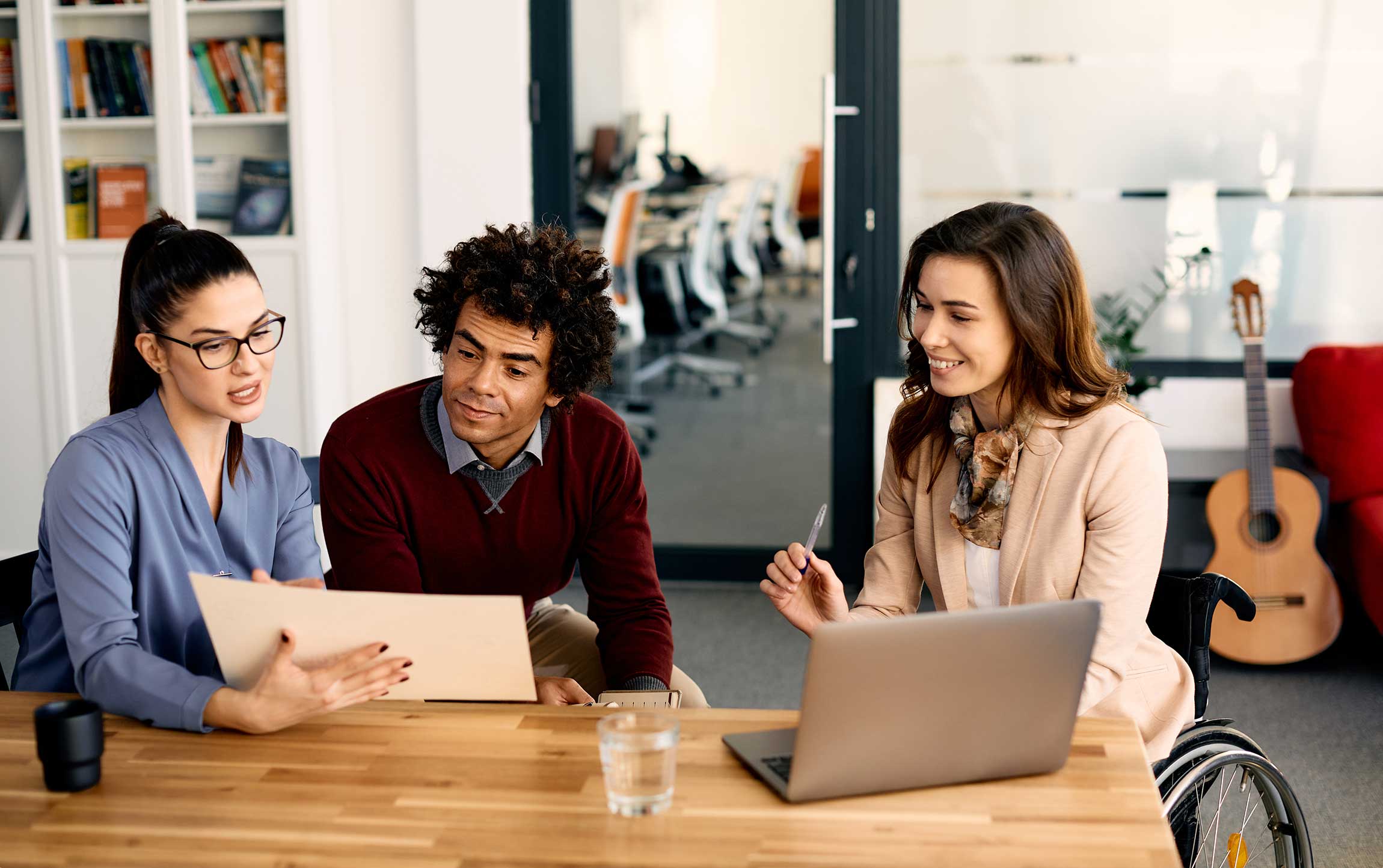 Business meeting in a modern office setting. On the left, a woman in a blue shirt is discussing a document with a man in a burgundy sweater. On the right, a woman in a beige blazer is working on a laptop and engaging in a conversation, seated in a wheelchair.