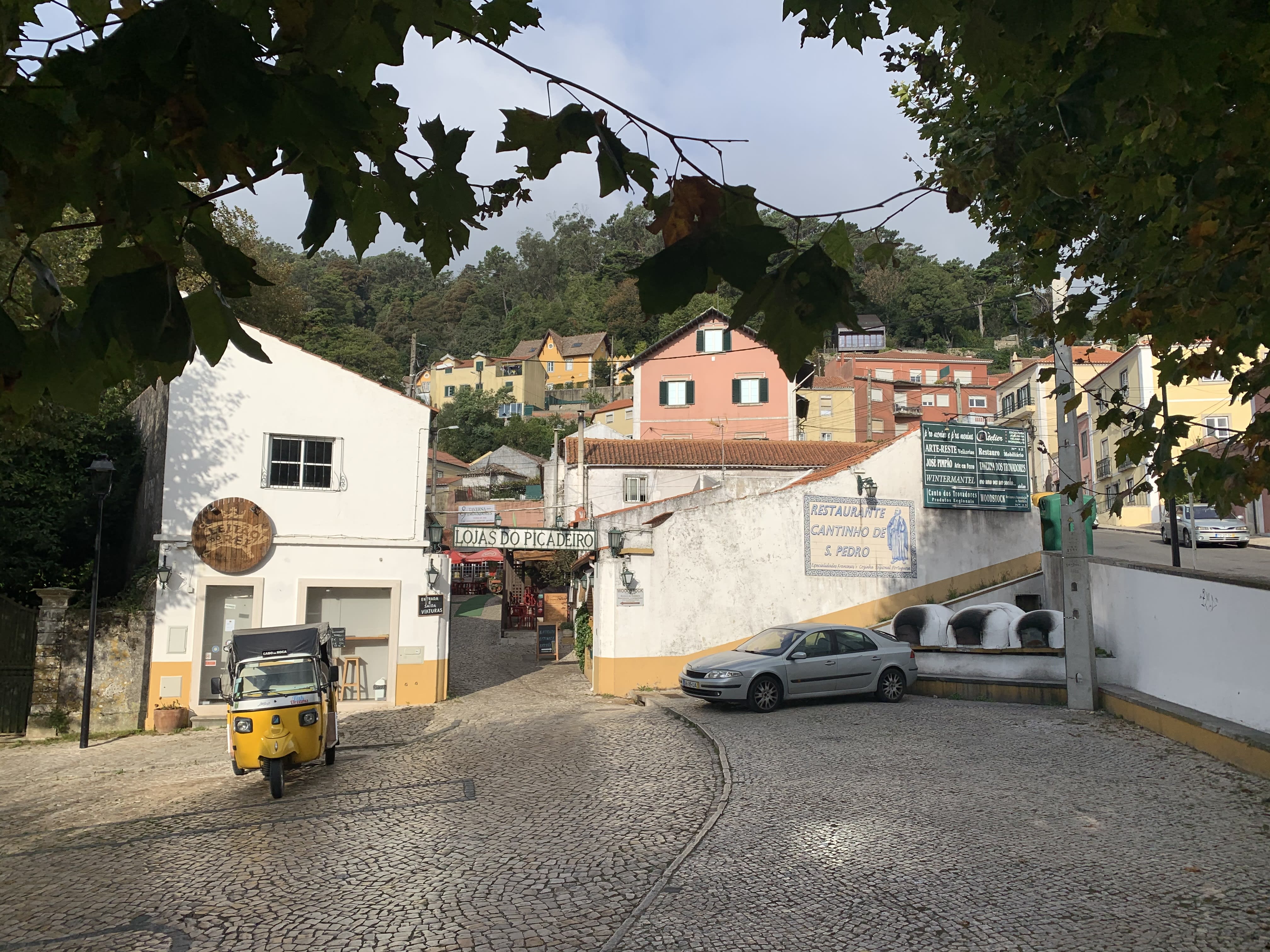 A quaint cobblestone street in a village near Sintra, Portugal, with colorful buildings, a small yellow vehicle, and signs indicating shops and restaurants lining the narrow pathway.