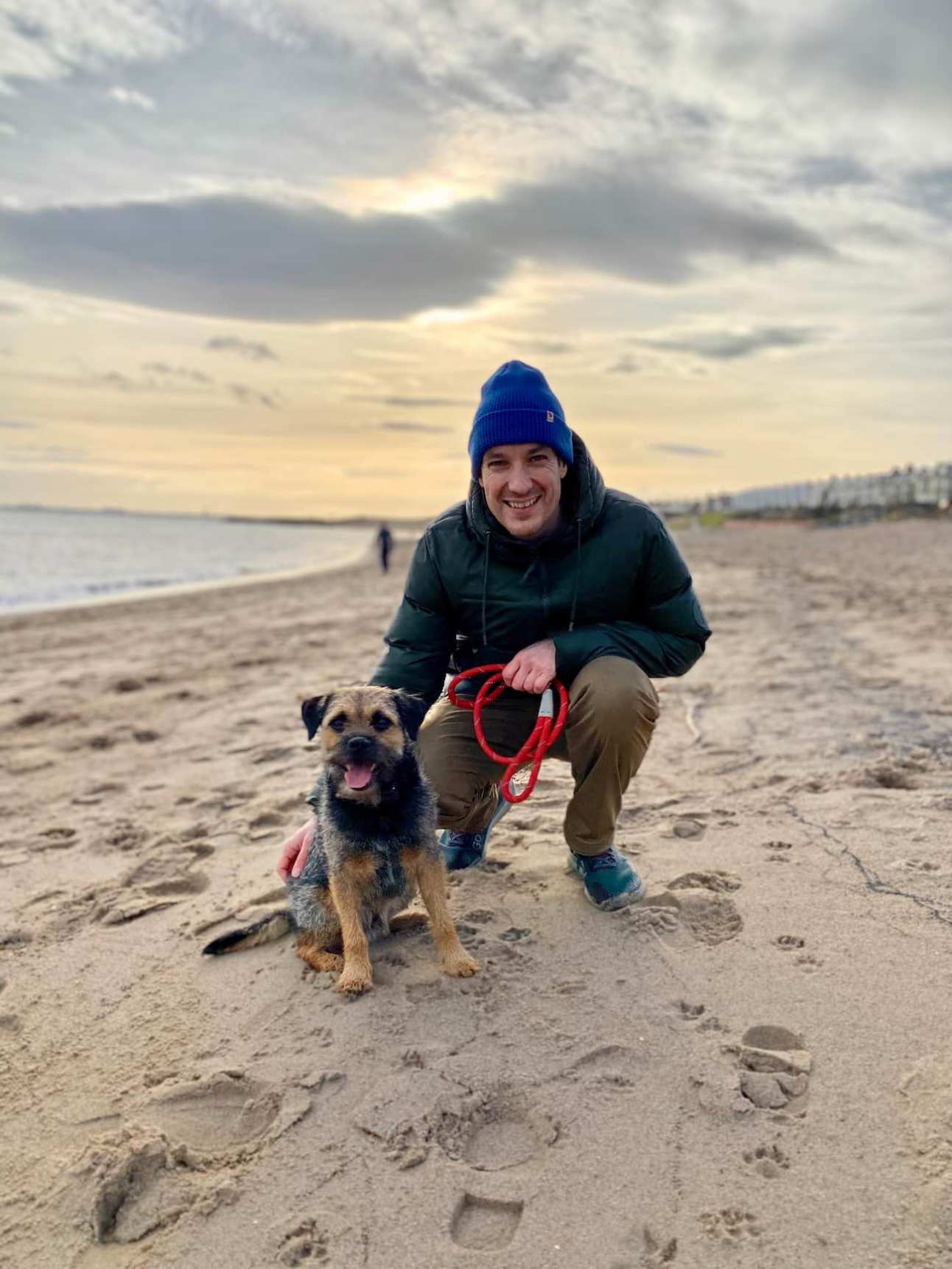 A smiling man wearing a green jacket and blue beanie hat crouches beside his happy border terrier dog on the beach